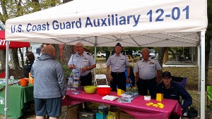 The lettering "U.S. Coast Guard Auxiliary 12-01" is visible on the canopy at the top of the photo, and Steve Stranevas, Joe Harris, Jay Price (standing), and Garrett Stengel (sitting) are facing out from behind the display table underneath the canopy.