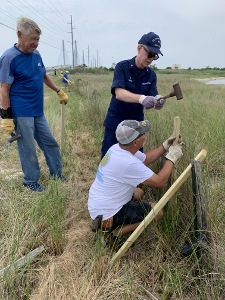 Bruce White, in ODU, pounds in a fence stake being held by a volunteer who is kneeling as a second volunteer stands behind them