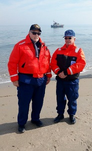 Fran, on left, and John face the camera wearing orange float coats with the ocean and a boat behind them