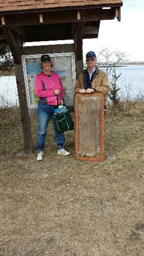 Barbara (at left) and Bruce White (in civilian clothers) stand at the kiosk at the entrance to the Burton Island Trail. Barb is holding a cloth bag fuill of trash and Bruce has the section of fiberglass in front of him, whiich comes up to his waist.