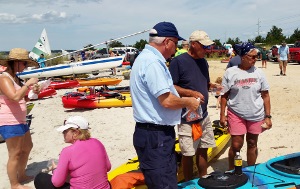 Denny Durgard prepares to take out a safety decal to affix to an inspected kayak. Other kayaks as well as sailboats are visible behind him on the shoreline