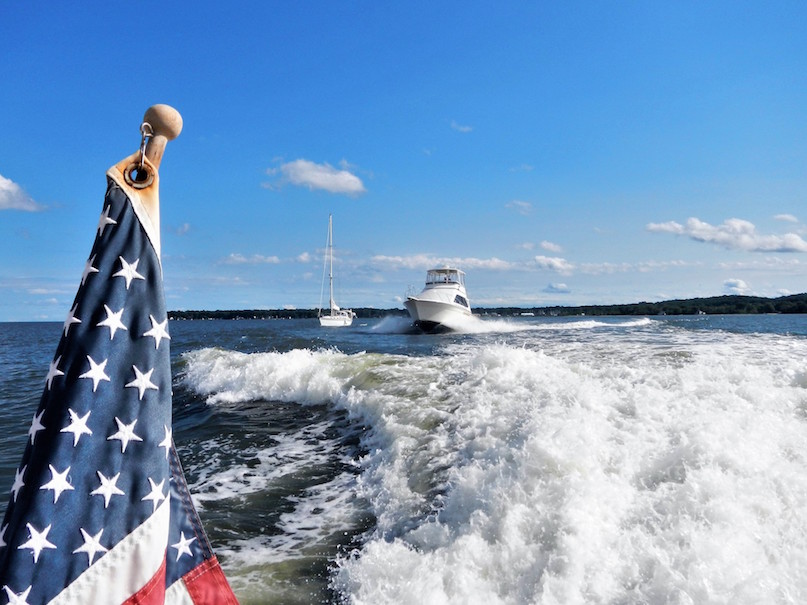 Picture taken from power boat. American flag in foreground. Power boat and sail boat in background.