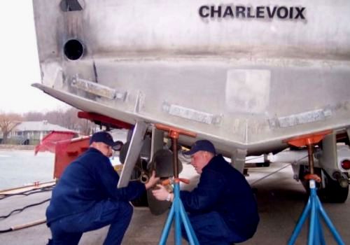 Two men in Coast Guard Uniforms working under a grey metal boat which bears the name Charlevoix
