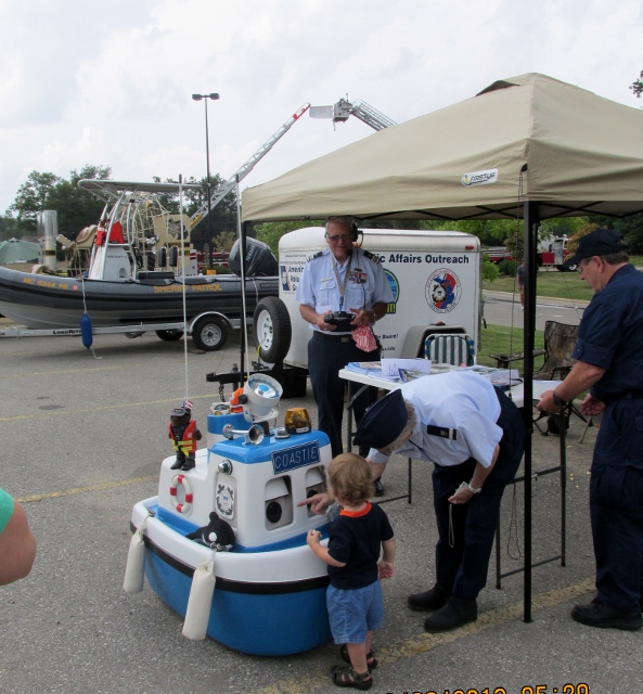Scene in a parking lot area with members of the Auxiliary incoast Guard Unifroms with the small Coast Guard Boat Robot and a young boy 