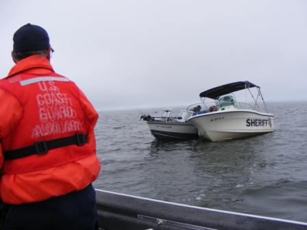 A man in a Coast Guard Auxiliary Life Vest looks on as a white sheriff's boat assists people our of a small recreational Boat 