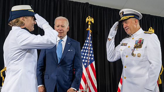 Adm. Fagan and Adm. Schultz, both wearing dress white uniforms, salute each other; President Biden watches; he is wearing a dark blue suit, a light blue tie, and white shirt