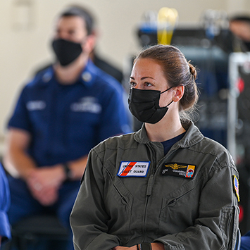 A seated service member wears a mask and a flight suit and looks off camera