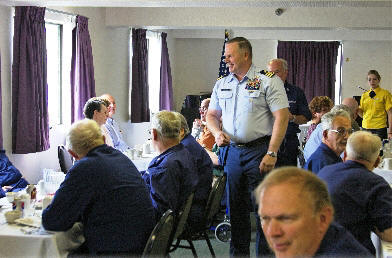 Several People in Coast Guard Uniforms at a banquet table. Standing next to the table is a man in a Coast Guard Officers Uniform.  All of them are laughing 
