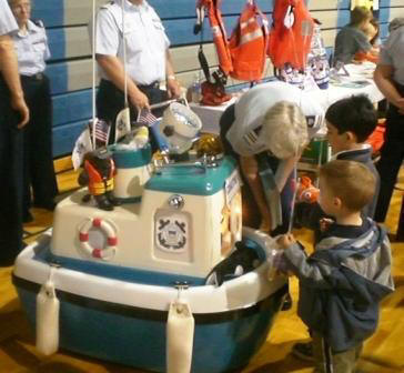 Several people at a safety booth. Children surrounding a blue and white robot which looks like a Coast Guard Boat 